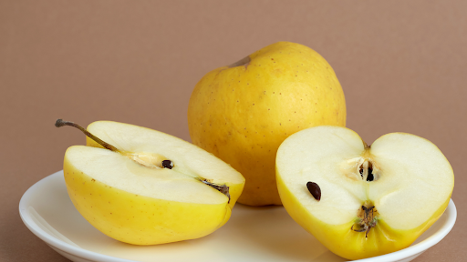 Two golden apples lie on a white plate. One of the apples is whole, the other is cut in half. The inside of the cut apples appears white, juicy and crisp. Seeds from the sliced apple are visible.